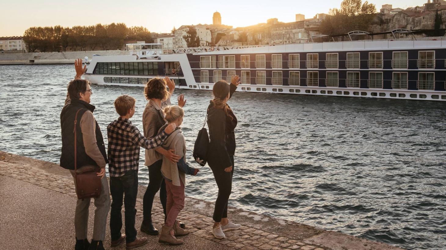 A group of 5 people stand on the shore waving at a passing river boat ship